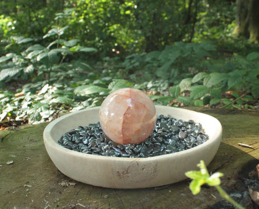 HÄMATIT Mini - Trommelsteine zur Reinigung von Heilsteinen in einer Holzschale auf einem Baumstamm mit einer Hämatoid Kugel in oben auf den Trommelsteinen und im Hintergrund Wad.