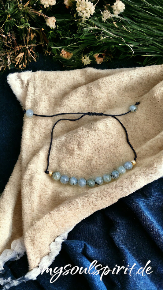 Das Labradorit Heilstein-Armband mit 10 grau-blauen Labradorit-Perlen in der Mitte, fixiert mit zwei Goldperlen, am Ende beiden Zugschnüre jeweils eine Perle. Das Armband ist größenverstellbar. Das Armband liegt zur Dekoration auf einem gelblichen Tuch.