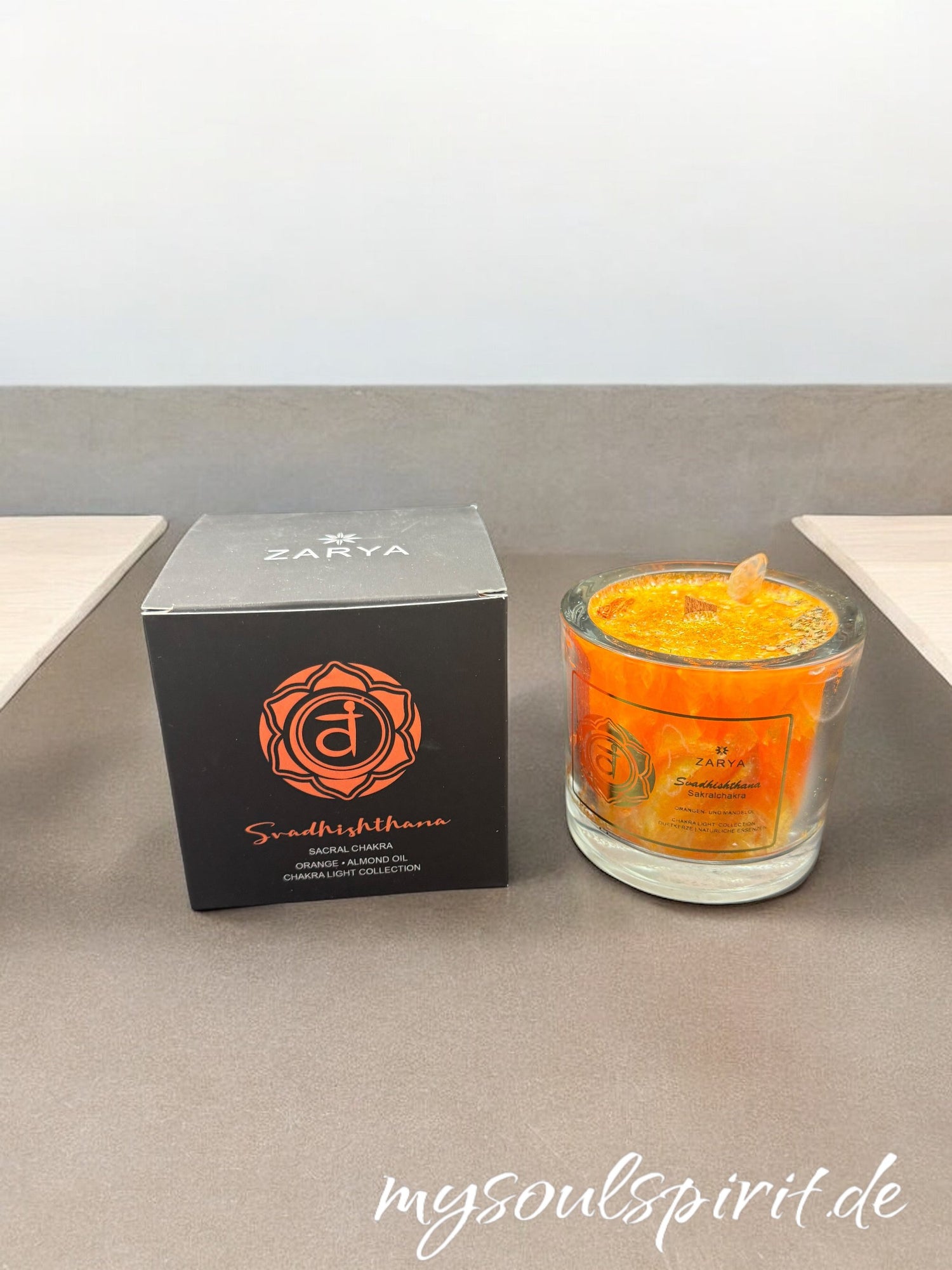 Orangene Kerze im Glas mit Heilsteinen Citrin und Orangencalcit,  sowie Orangenblüte als Duft. Daneben steht die quadratische Verpackung in schwarz mit orangener Schrift und dem Sakral-Chakra Symbol